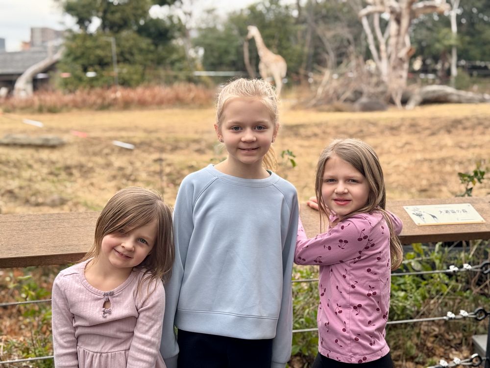 Lily, Harper, and Cora standing at a railing at Tennoji Zoo in Osaka with a giraffe visible in the enclosure behind them