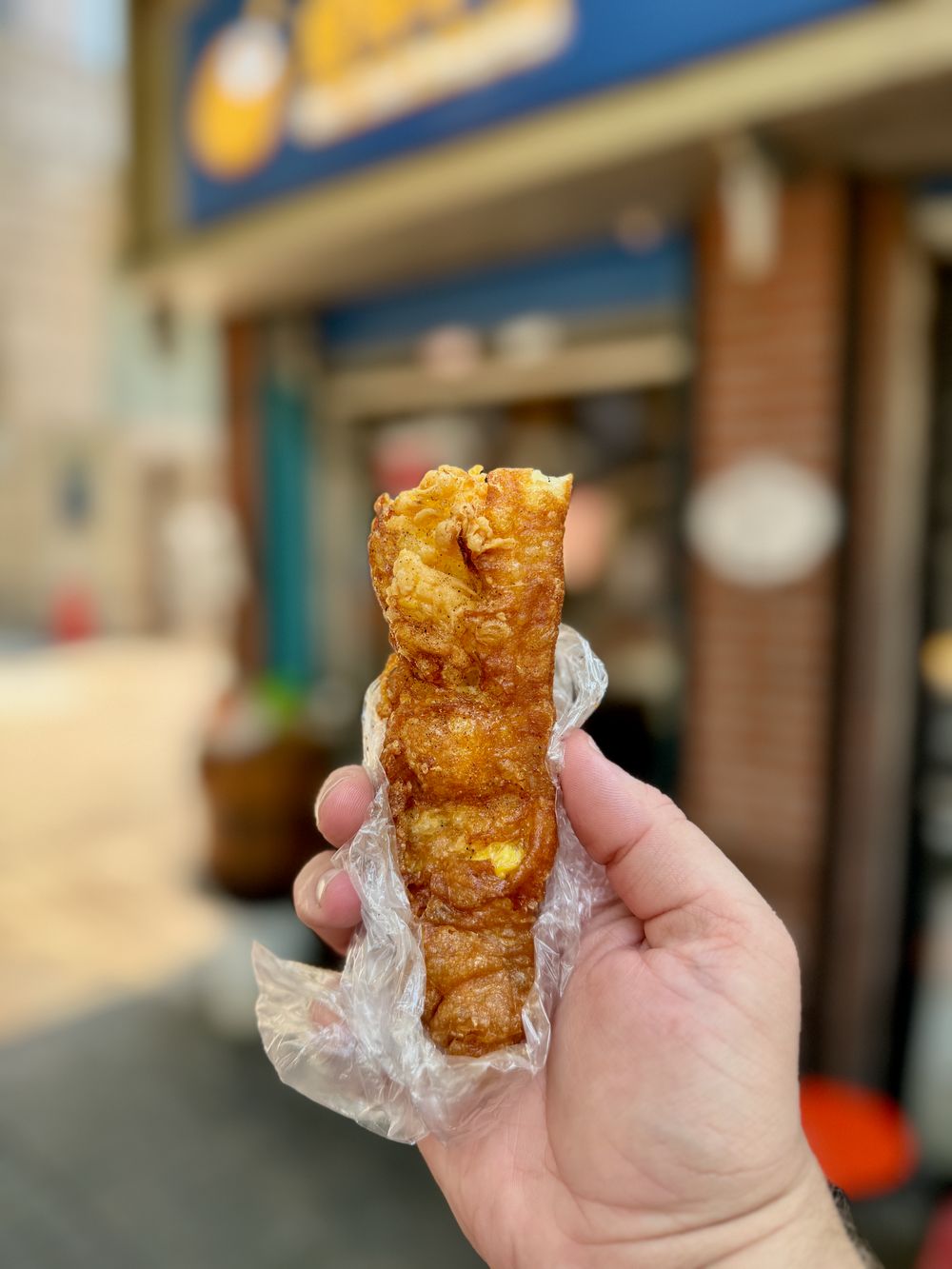 Hand holding a golden fried egg roll wrapped in plastic in front of a shop in Jing'an District Shanghai