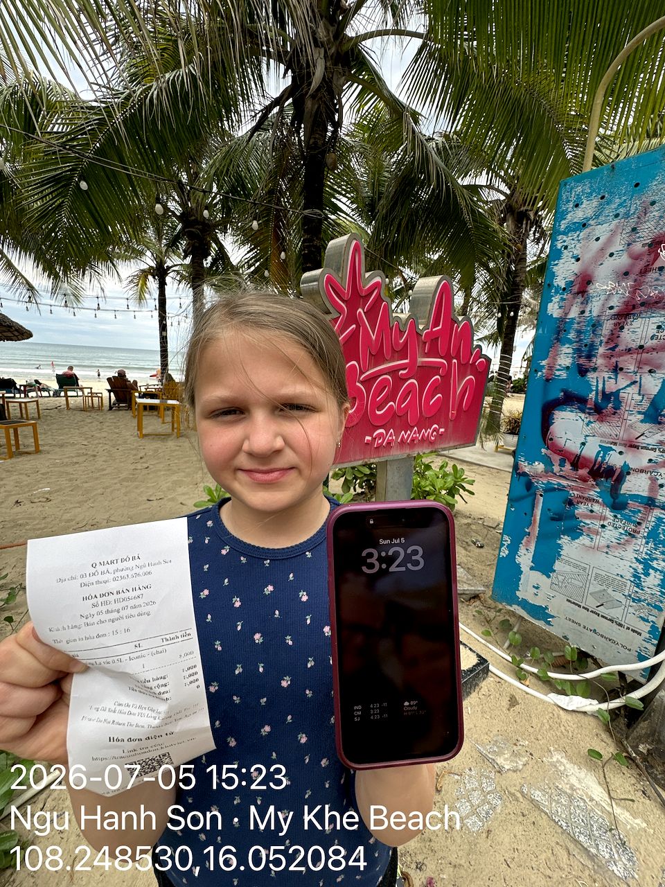 Cora standing in front of the pink My An Beach sign in Da Nang holding a Vietnamese receipt and a phone, with the beach behind her