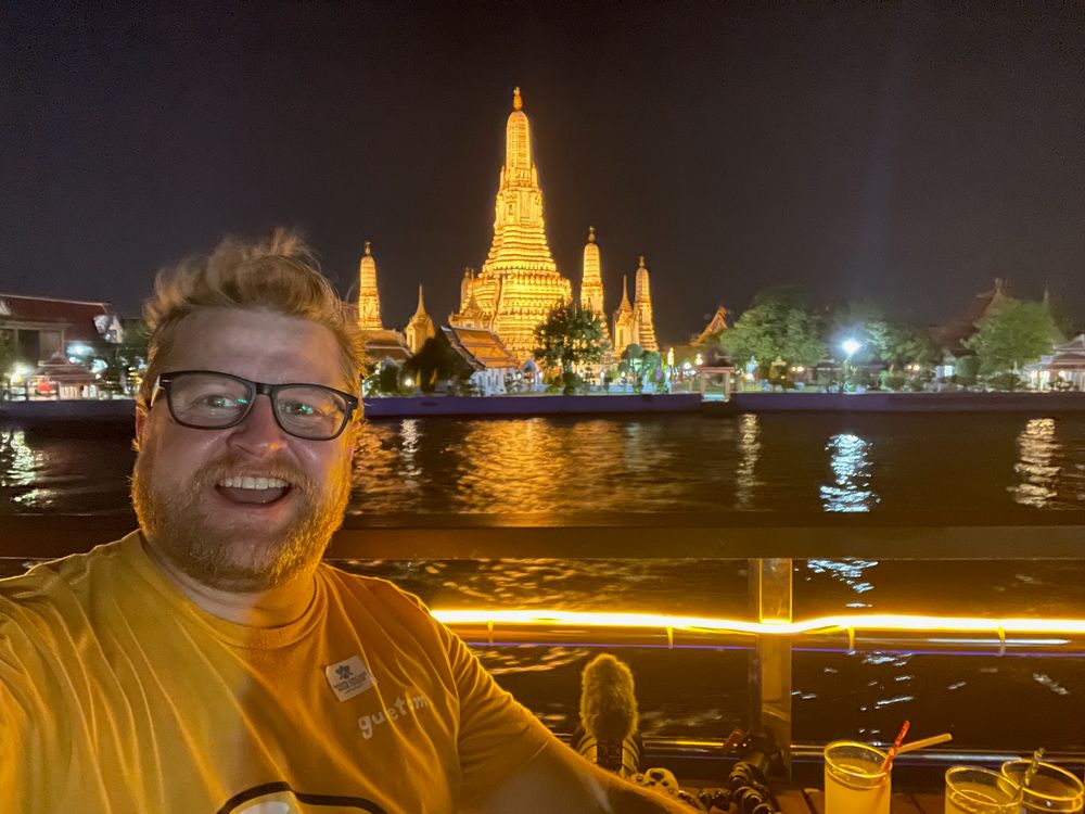 Adam smiling in a selfie on a river boat at night with the illuminated spires of Wat Arun reflected in the Chao Phraya River behind him.