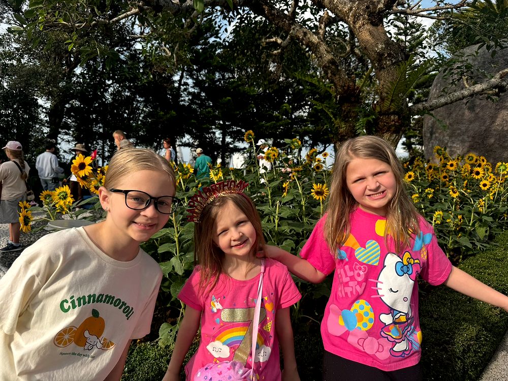 Three young girls standing and smiling in front of a field of sunflowers at Ba Na Hills, the middle girl wearing a sparkly gold Happy Birthday crown