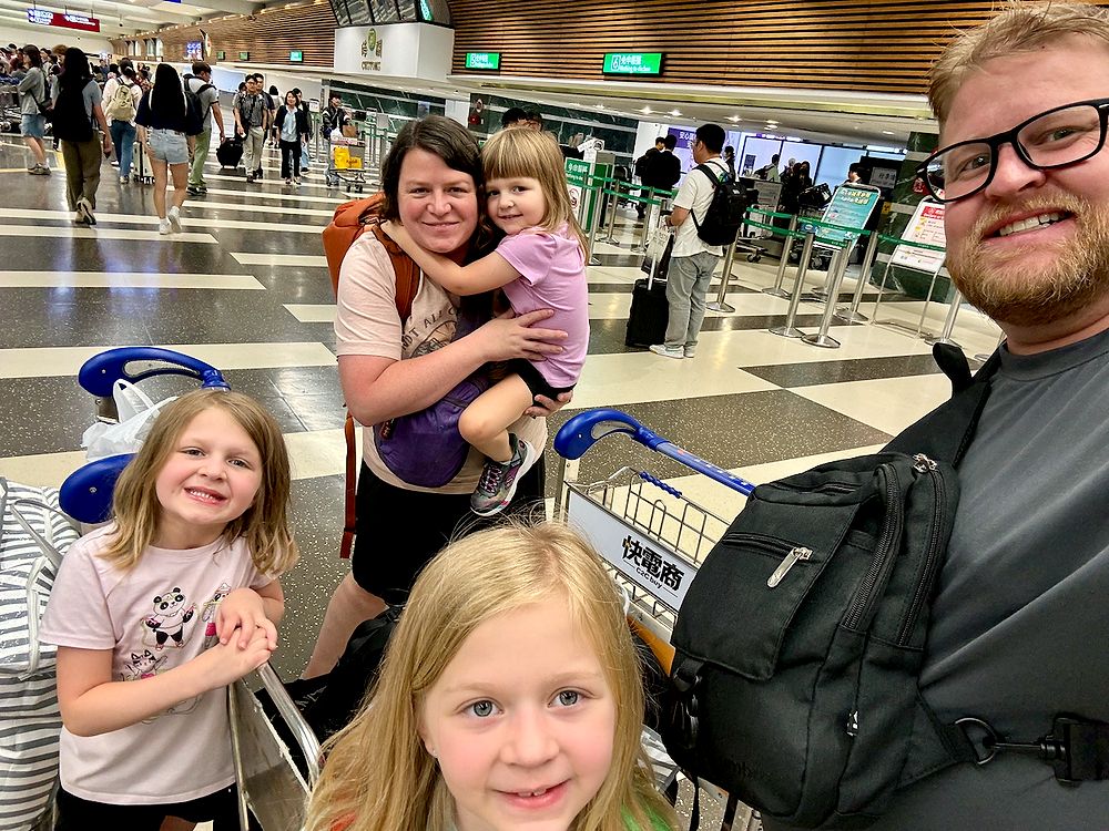 The Clarkson family at Taoyuan International Airport with a loaded luggage cart, Harper in Lindsay's arms, all five smiling on arrival in Taiwan