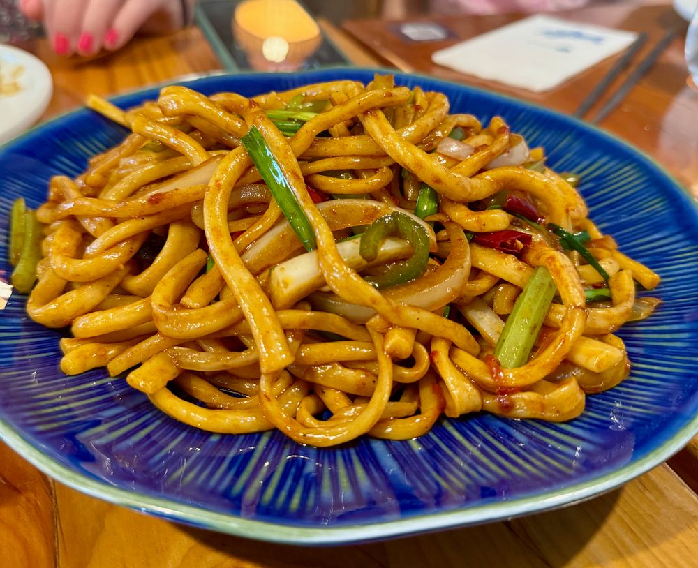 Large blue ceramic plate of thick spicy cumin hand-pulled noodles in chili oil with green onions and dried red chilies, Jing'an Shanghai fall 2025
