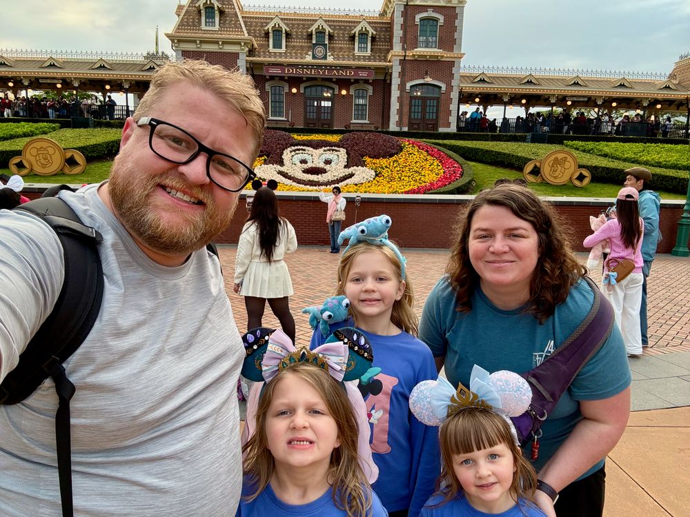 Family selfie in Minnie ears at the Hong Kong Disneyland entrance with the Mickey floral display behind them.
