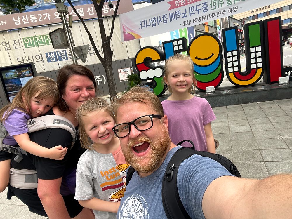 Family selfie in front of the large colorful Seoul sign in Myeongdong, with Adam in the foreground, Lindsay carrying Harper, and Lily and Cora smiling behind