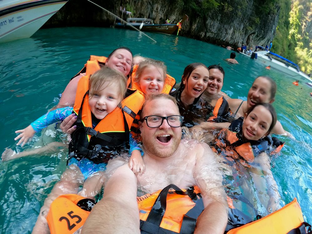 Adam takes a GoPro selfie in turquoise water surrounded by the whole family in orange life jackets, with longtail boats and limestone cliffs visible in the background.