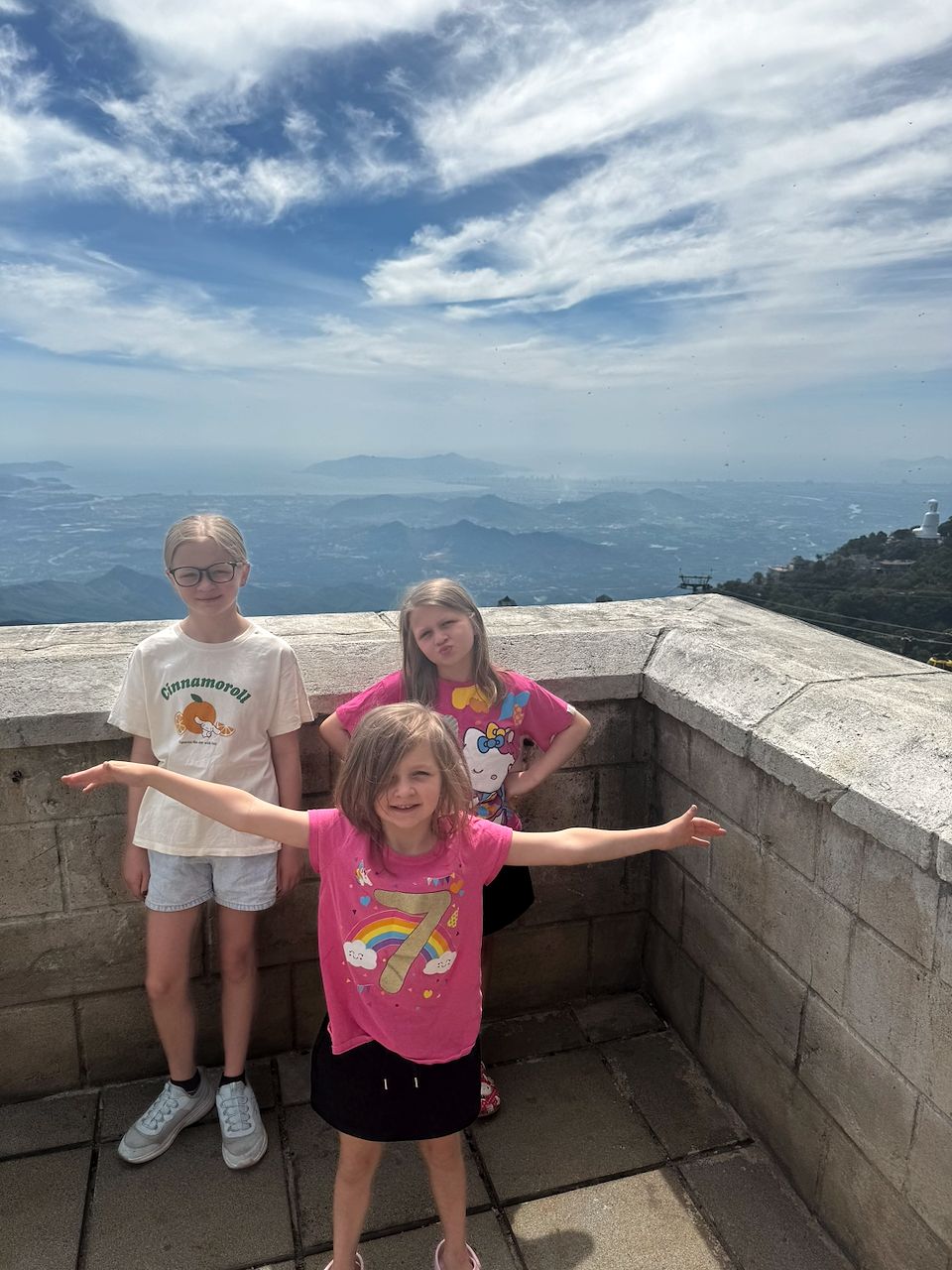 Three girls pose on a stone parapet at the top of Ba Na Hills, with a sweeping hazy mountain and coastal view behind them; the youngest in a pink birthday shirt spreads her arms wide while the older two stand on either side.