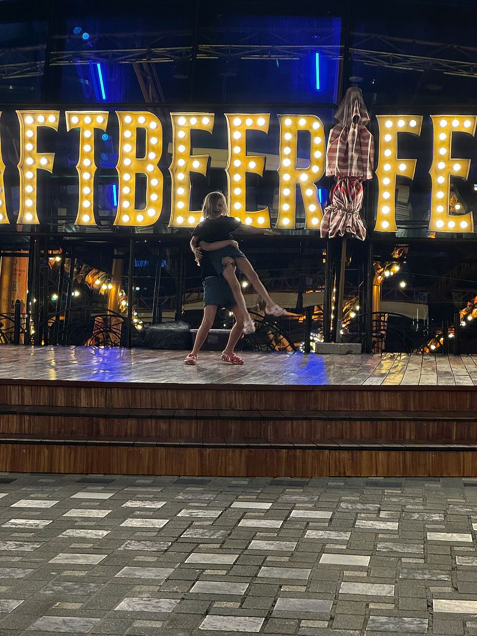 Two children dancing together on a wooden stage in front of large illuminated marquee letters spelling Craft Beer Fest at an outdoor venue at night.