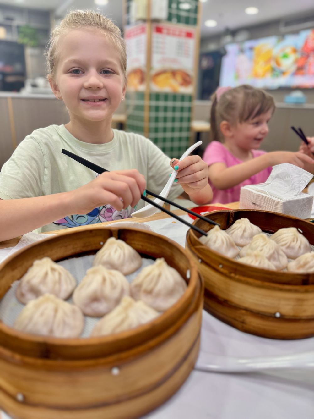 Harper smiling with chopsticks over a basket of xiaolongbao at a restaurant in Xuhui District, Shanghai, Lily eating in the background, fall 2025