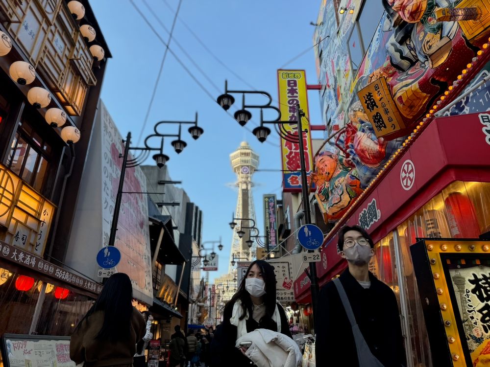 A busy Shinsekai shopping street in Osaka lined with lanterns and signs, with Tsutenkaku tower at the far end under a blue sky