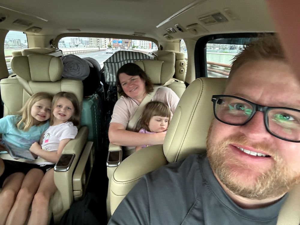 Adam taking a selfie from the front seat of an airport taxi in Tokyo with Lindsay and three girls in the back, July 2024