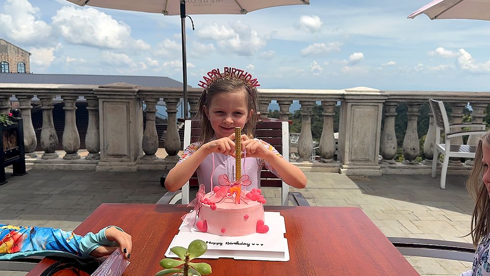 A young girl wearing a glittery Happy Birthday crown sits at an outdoor table on a stone terrace, hands gripping a tall birthday candle on a pink heart-decorated cake, with a blue sky and mountain views behind her.