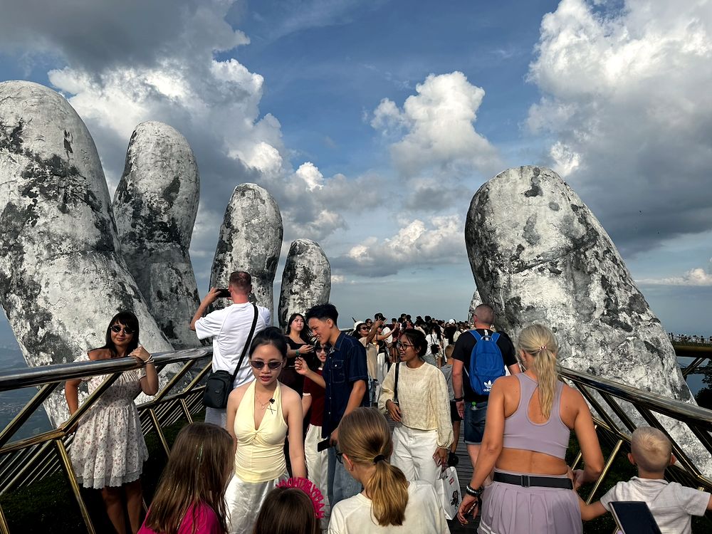 A dense crowd of tourists walks along the Golden Bridge at Ba Na Hills, flanked by two massive weathered stone hands rising against a partly cloudy blue sky.