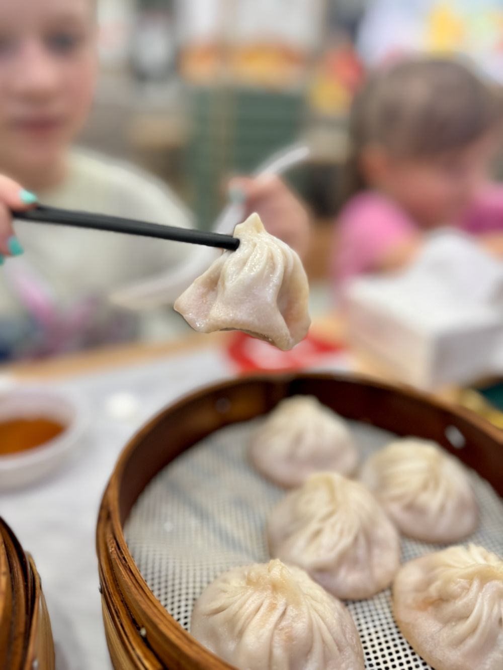 A xiao long bao held up with black chopsticks over a bamboo steamer basket, Xuhui District Shanghai fall 2025