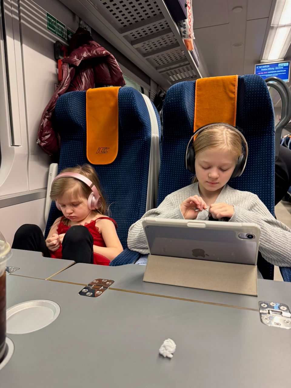 Harper and Lily sitting side by side on a Polish intercity train wearing headphones, with an iPad propped on the tray table