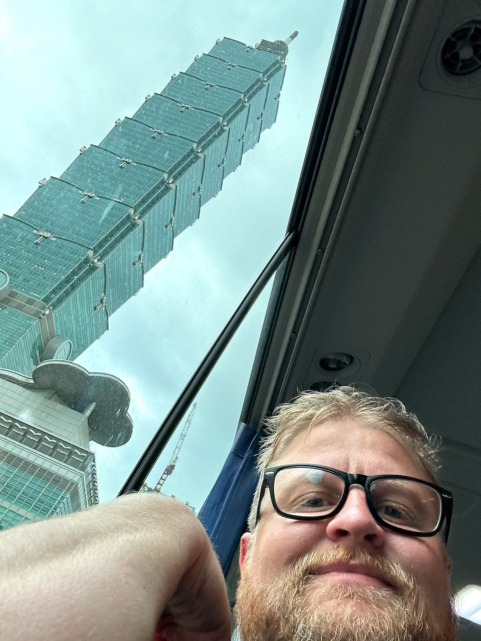 Adam taking a selfie on a bus with Taipei 101 visible through the rain-streaked window behind him