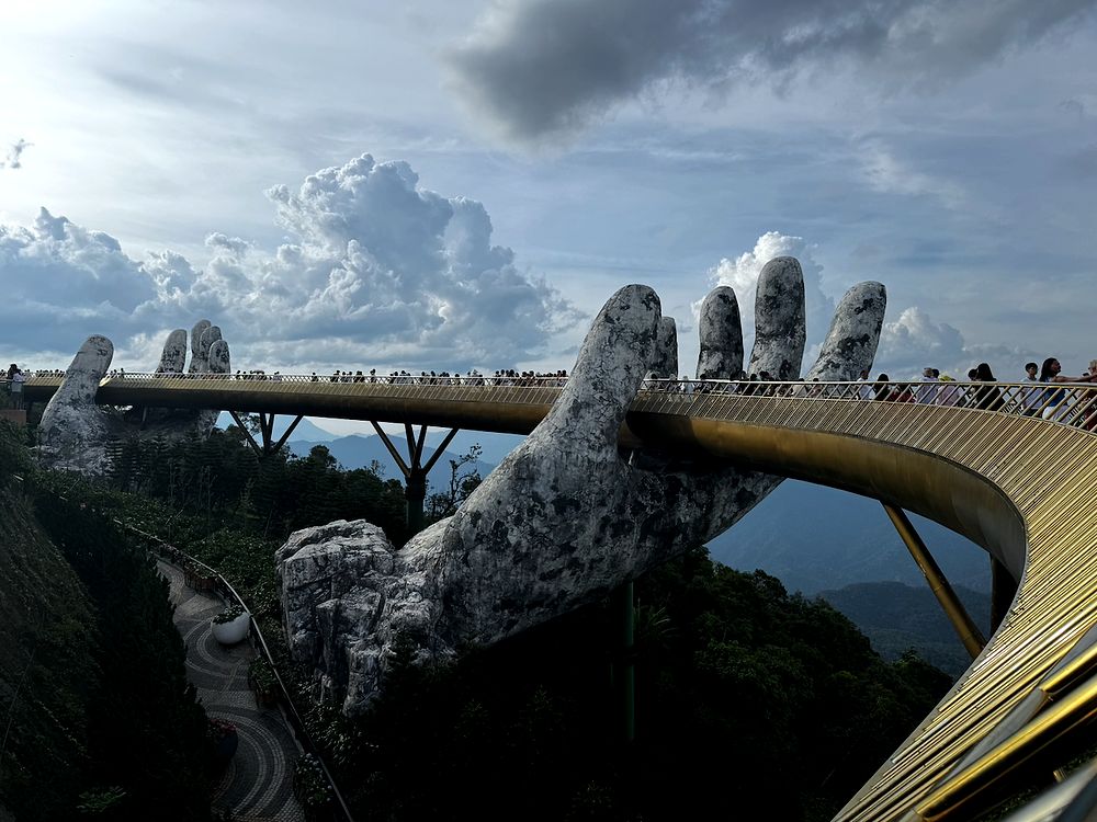 The Golden Bridge at Ba Na Hills, a golden walkway held up by two giant weathered stone hands rising from the forested mountainside under a dramatic sky