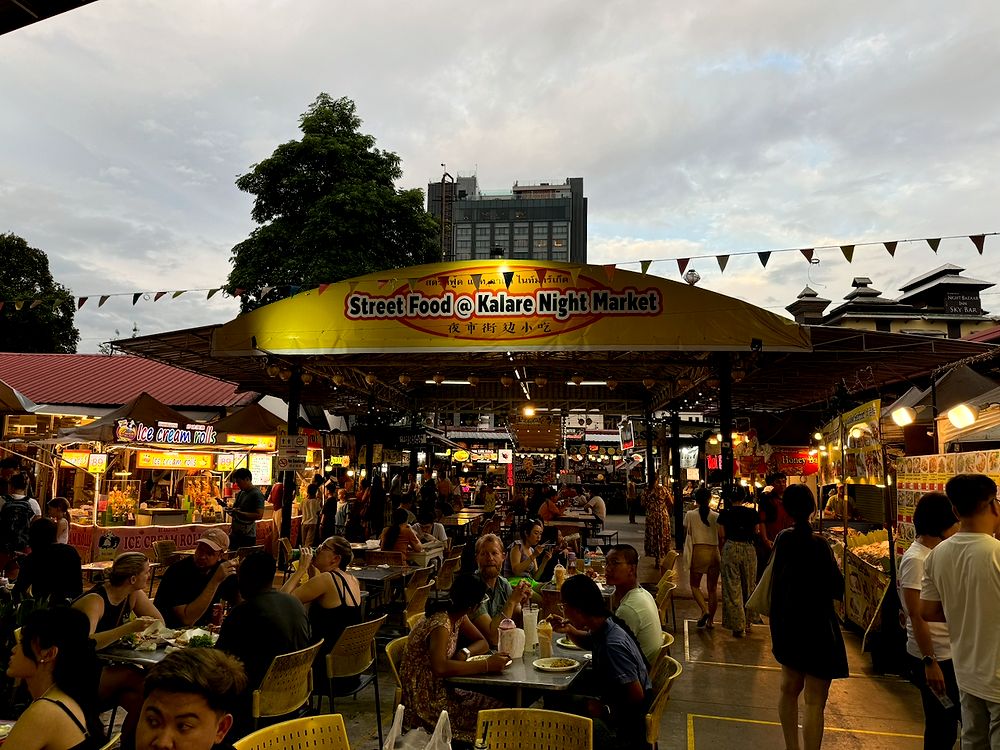 A wide shot of the Kalare Night Market in Chiang Mai at dusk, with a large yellow sign reading Street Food at Kalare Night Market, crowded tables of diners, surrounding food stalls lit up, and an overcast evening sky above.