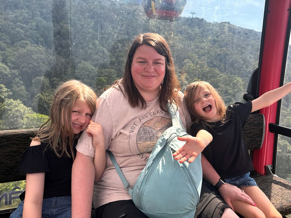 A woman sits inside a cable car gondola with a girl leaning into her on the left and a younger girl with arms spread wide laughing on the right, forested mountains and another gondola visible through the glass behind them.