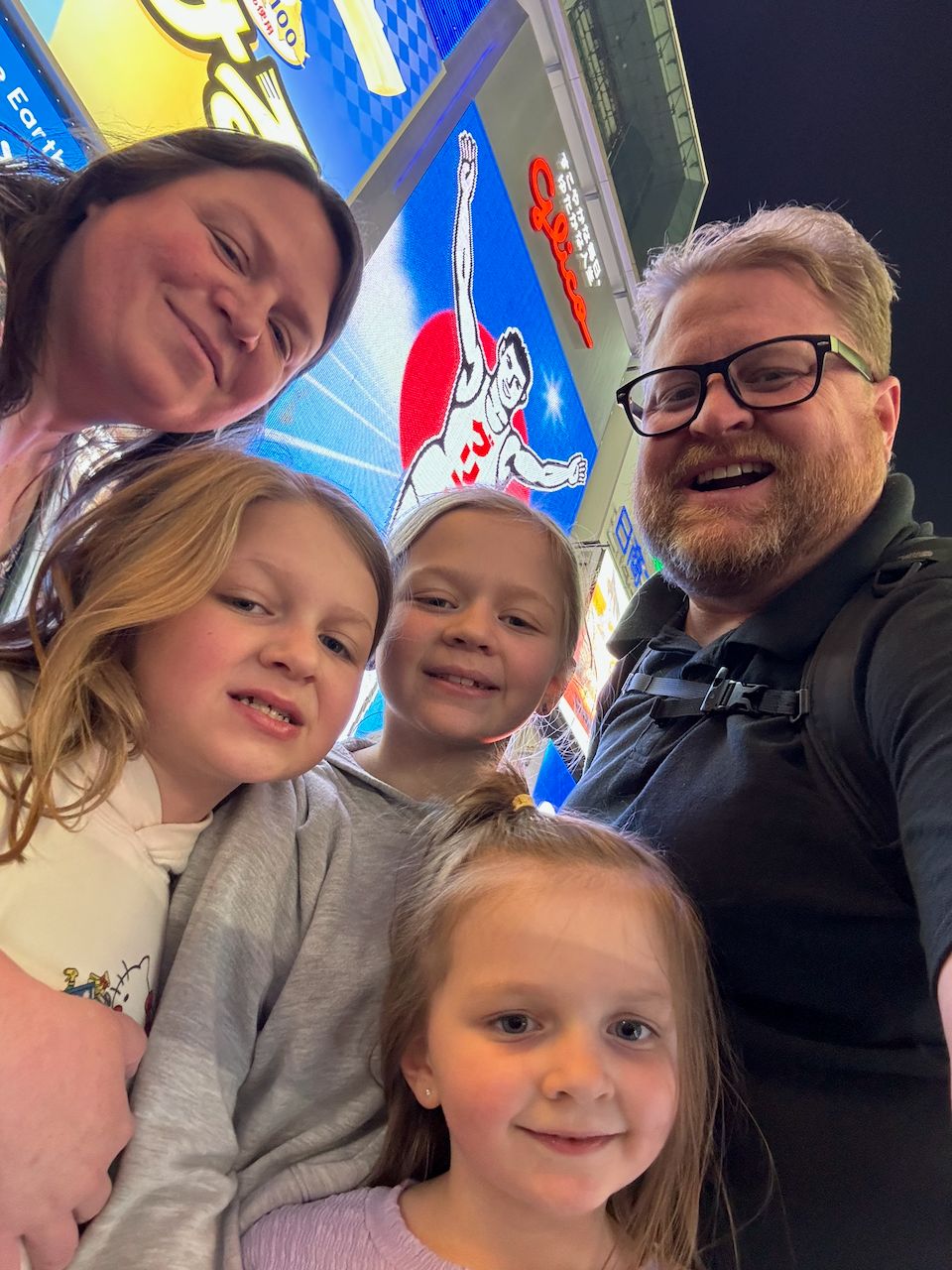A family selfie of two adults and three young girls in front of the illuminated Glico Man sign on a neon-lit Dotonbori bridge at night in Osaka