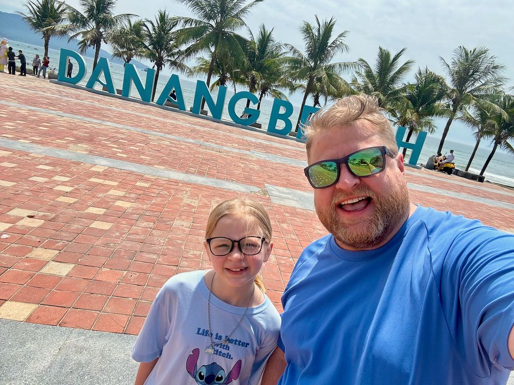 Adam and Lily take a selfie in front of the large teal Da Nang Beach sign on a sunny day